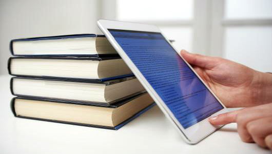 Image of hands holding an iPad leaning against a stack of books. Image is for decorative purposes only. It is not informational.
